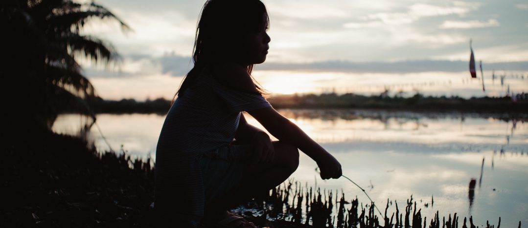 The silhouette of a child crouching along a waterfront at sunset.