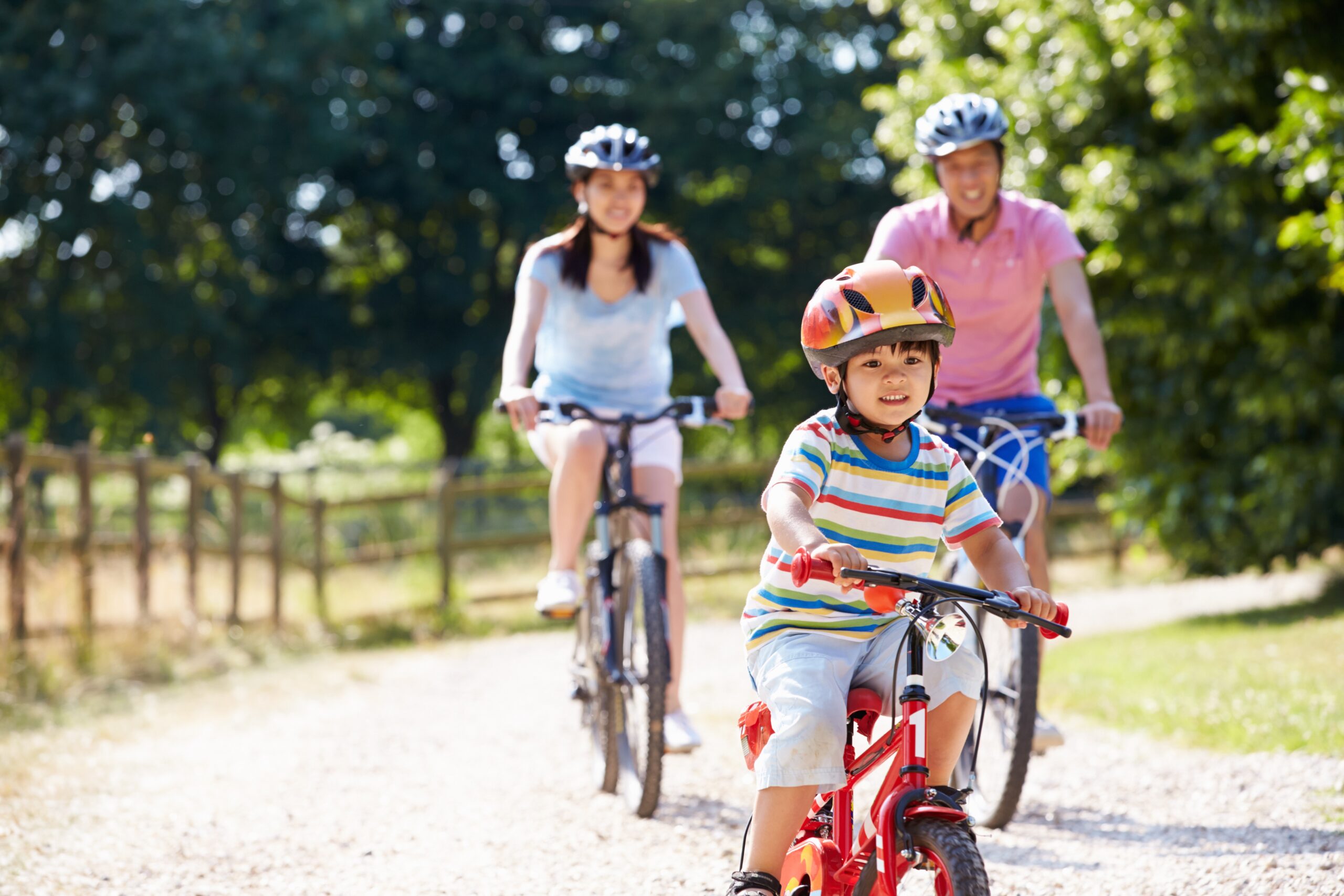 Two adults and a child ride their bikes on a gravel path through trees.