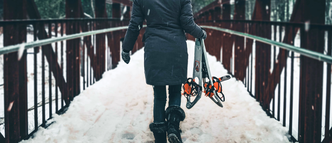 woman walking across a snowy bridge wearing a big winter coat, snow boots, and carrying snowshoes