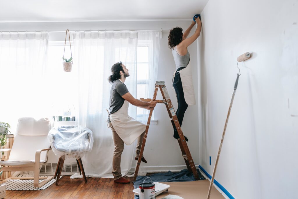 A man holds a ladder while a woman stands on it and paints in a home.