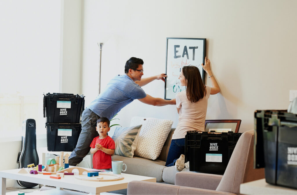 Two parents moving into a home hanging a picture on the wall, while their child plays with toys on a coffee table nearby.