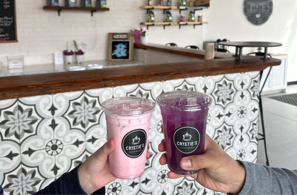 Two hands cheersing pink and purple seasonal iced drinks at Crystie's Coffee.