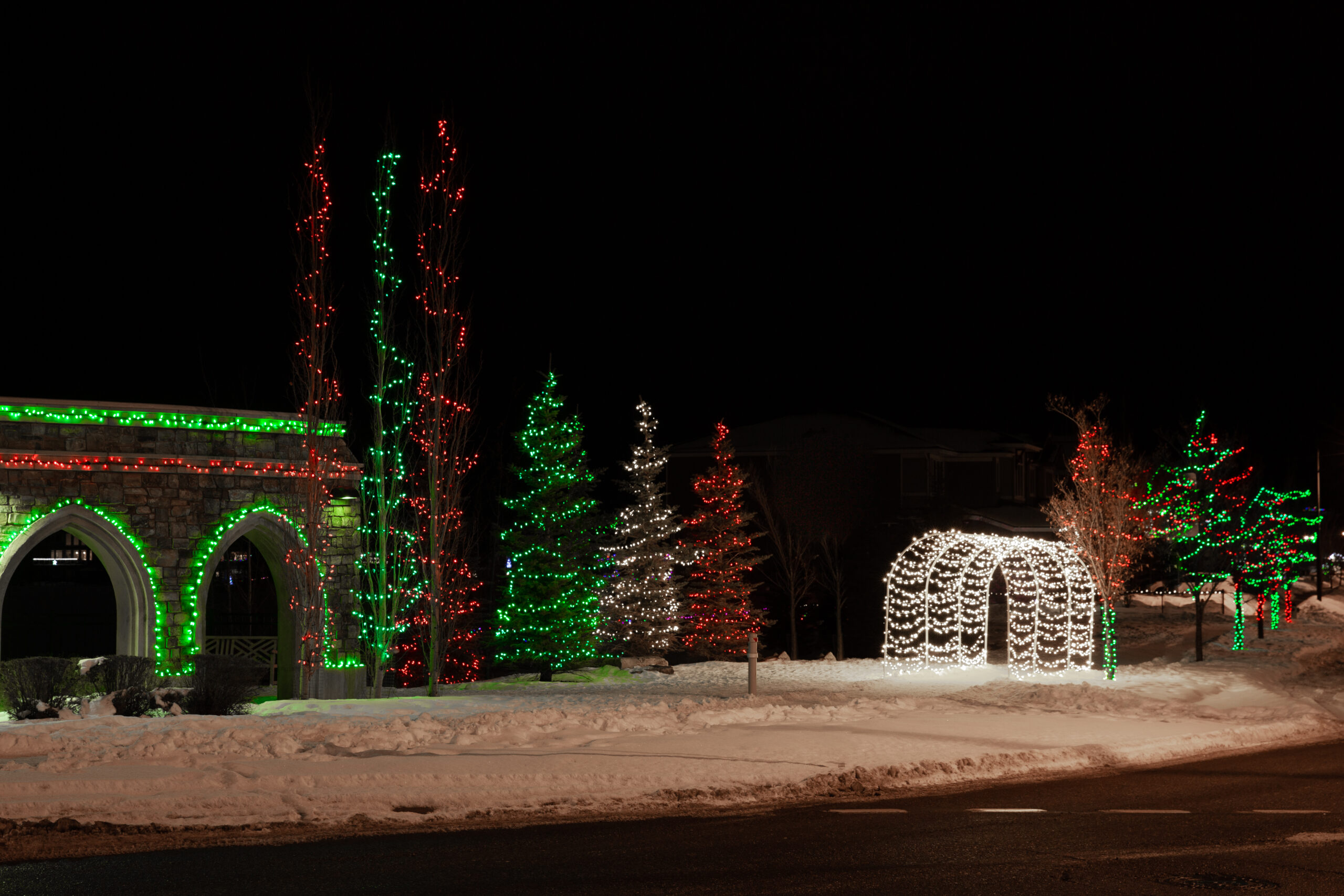Christmas lights decorating a stone archway, several trees and and a pedestrian arch in the community of Legacy.